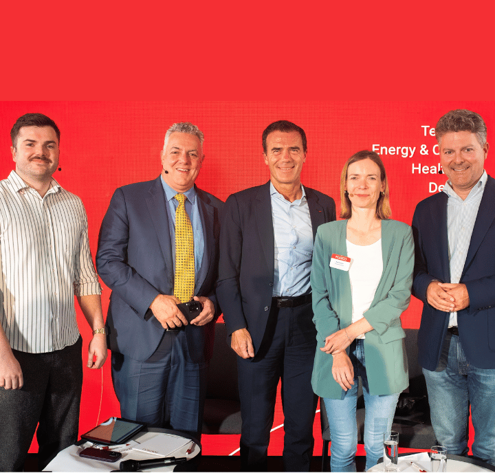 Five people stand side by side, smiling at the camera, against a bright red background with faint white text. The group consists of three men in suits and two casually dressed individuals, a man and a woman. Tablets and microphones rest on a table in front of them.