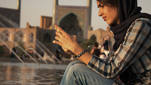 A woman wearing a plaid shirt and a dark headscarf sits by a pool, smiling while using her smartphone. The background shows a historic building with arched entryways and fountains, likely in a Middle Eastern or Central Asian setting.