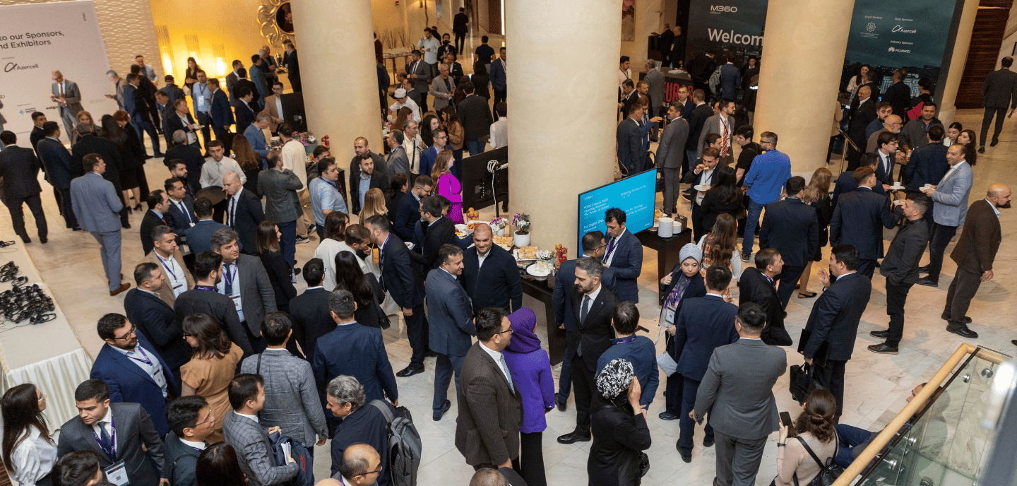 A large crowd of people in formal attire mingle in a bright lobby with tall columns. Booths, banners, and monitors advertising a conference or expo are visible. Some people chat, others walk or look at exhibits. The atmosphere is lively and professional.
