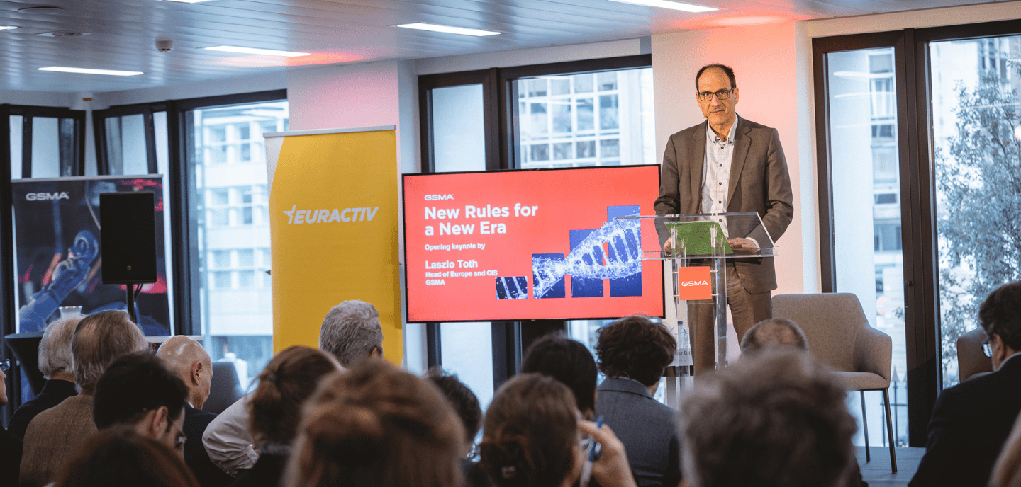 A man in a suit stands at a podium, speaking to an audience. Behind him, a screen displays “New Rules for a New Era” with DNA imagery. Banners for GSMA and EURACTIV are visible, and people sit attentively in a modern, well-lit conference room.