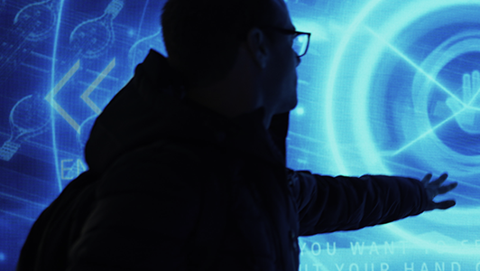 A person wearing glasses and a backpack extends their arm toward a glowing blue digital screen, which displays abstract shapes, circular patterns, and faint text. The scene has a futuristic and interactive technology vibe.