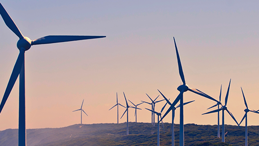 Several wind turbines stand on a hilly landscape under a clear sky at sunset. The turbines' blades are silhouetted against the soft, warm hues of the sky, suggesting renewable energy generation in a peaceful, natural setting.