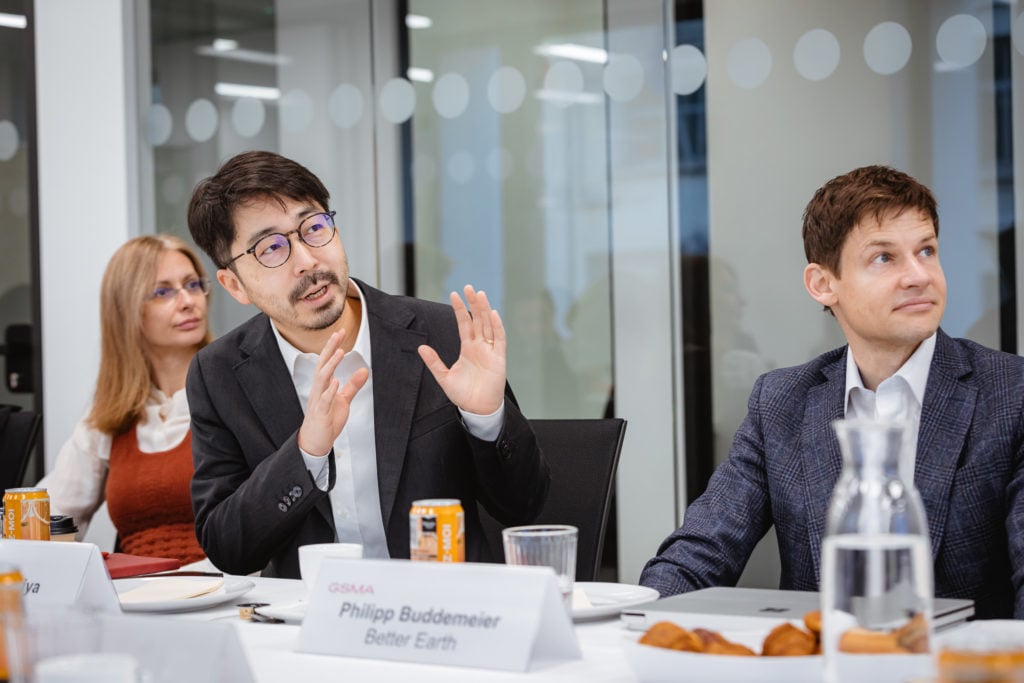 Three people sit at a conference table. The man in the center gestures while speaking, flanked by a woman on the left and a man on the right. Nameplates and a can of soda are visible on the table, alongside papers and glasses. The setting appears to be a modern office.