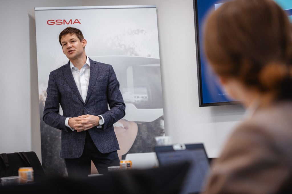 A man in a blue blazer and white shirt is speaking in front of a GSMA banner featuring a VR headset. He's standing in a conference room, addressing an audience. A woman with brown hair, seen from behind, is focused on him, with a laptop open in front of her.