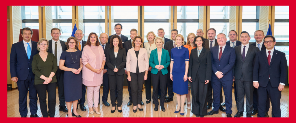 A group of 26 people, including men and women, are standing in rows posing for a photo in a brightly lit room with large windows. They are dressed in formal business attire. The setting suggests a professional or political gathering. The image is framed by a red border.
