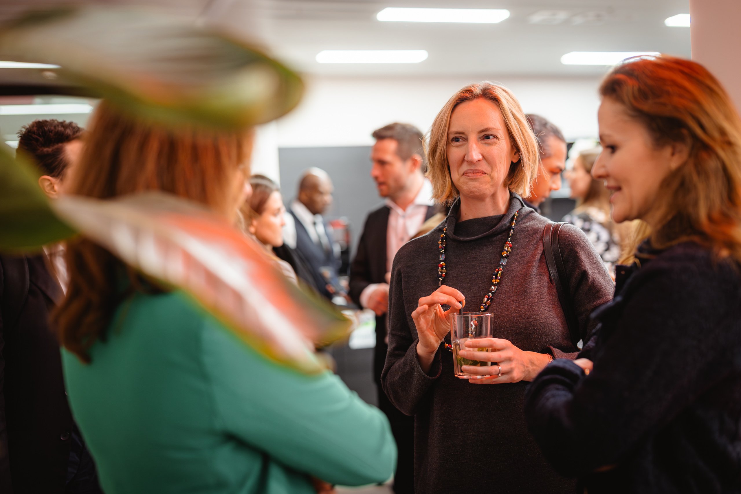 A social gathering in a brightly lit room with several people mingling. A woman in focus is holding a glass, smiling and wearing a dark sweater with a necklace. A blurred plant leaf is in the foreground on the left. Attendees in the background are engaged in conversation.