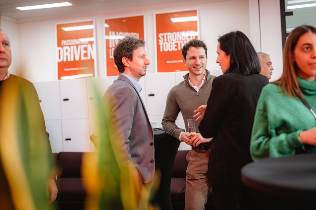 A group of people engages in conversation in a modern office with motivational posters on the walls. A man in a gray suit and a man in a green sweater are talking to a woman in black. There are black couches and a table nearby, with green plant leaves in the foreground.