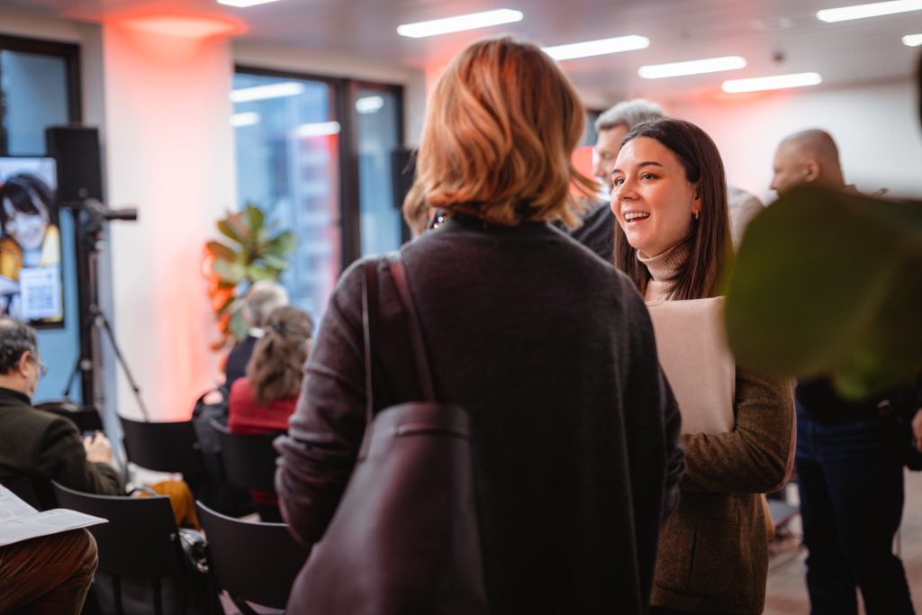 Two women converse at an indoor event. The woman on the right holds a folder and smiles, while the one on the left has her back to the camera. In the background, attendees sit facing a speaker, with large windows and orange lighting setting the ambiance.