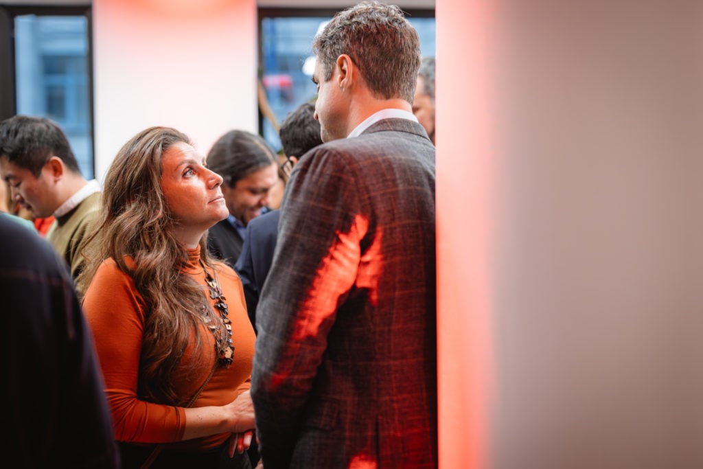 A woman in an orange top with long, wavy hair looks up at a man in a gray suit. They are indoors with a soft, warm light creating a red glow on the walls. Other people are blurred in the background, and the scene suggests a social gathering or event.