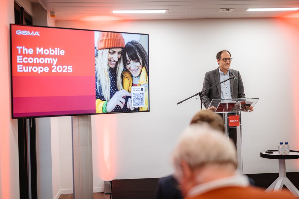 A man speaks at a podium in a conference room with an audience. A screen displays "GSMA The Mobile Economy Europe 2025" next to an image of two people looking at a smartphone. The room has white walls and red lighting. Bottles and a glass sit on a nearby small table.