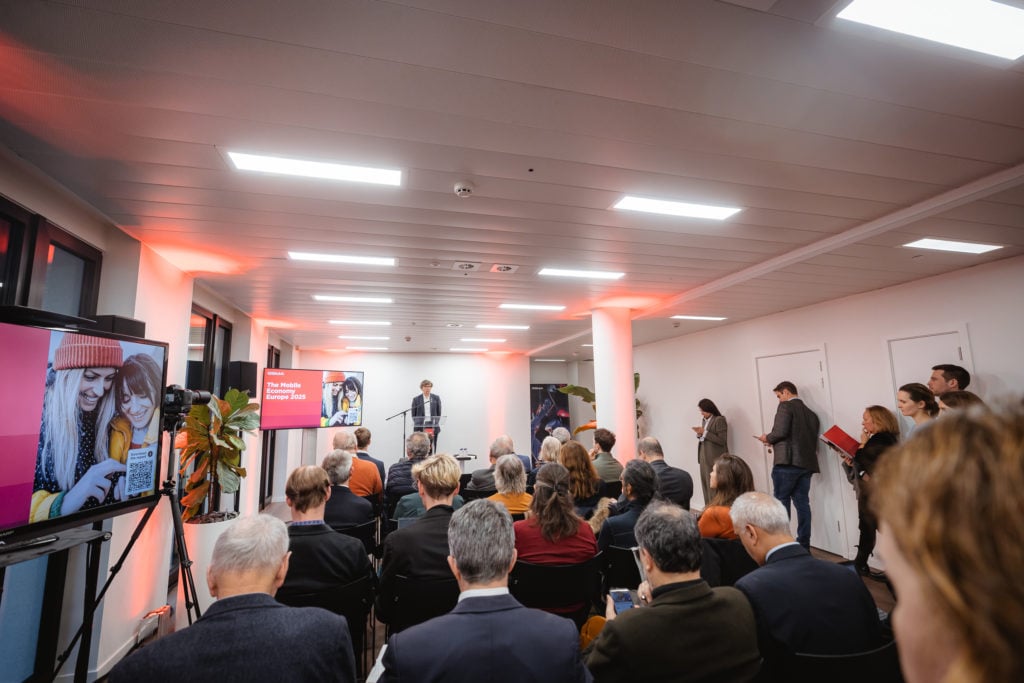 A speaker stands at a podium addressing a seated audience in a modern room, illuminated by bright lights. A large screen on the left displays a colorful image of two people smiling with a red overlay. Attendees are dressed in formal attire, and some are standing near the walls.