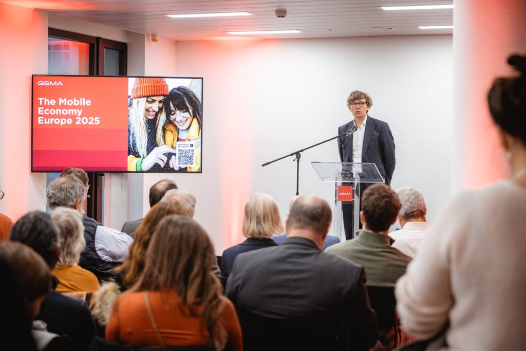 Man in suit speaks at a podium during an event. A slide on a large screen beside him reads "GSMA The Mobile Economy Europe 2025" with an image of two people looking at a tablet. The audience is seated, viewing the presentation. The room is brightly lit with red accents.