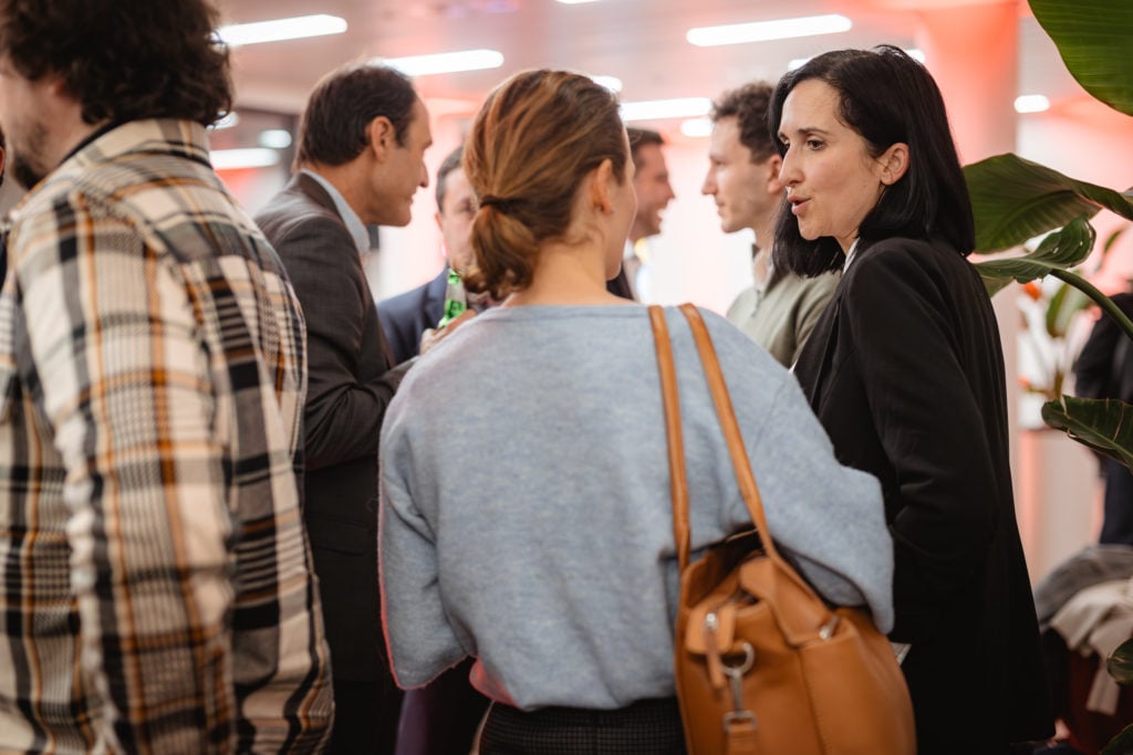 A group of people are engaged in conversation at a social event. A woman with dark hair is speaking to another individual with a purse slung on her shoulder, wearing a blue sweater. The background is softly lit in orange hues, and a man in a plaid shirt is visible in the foreground.