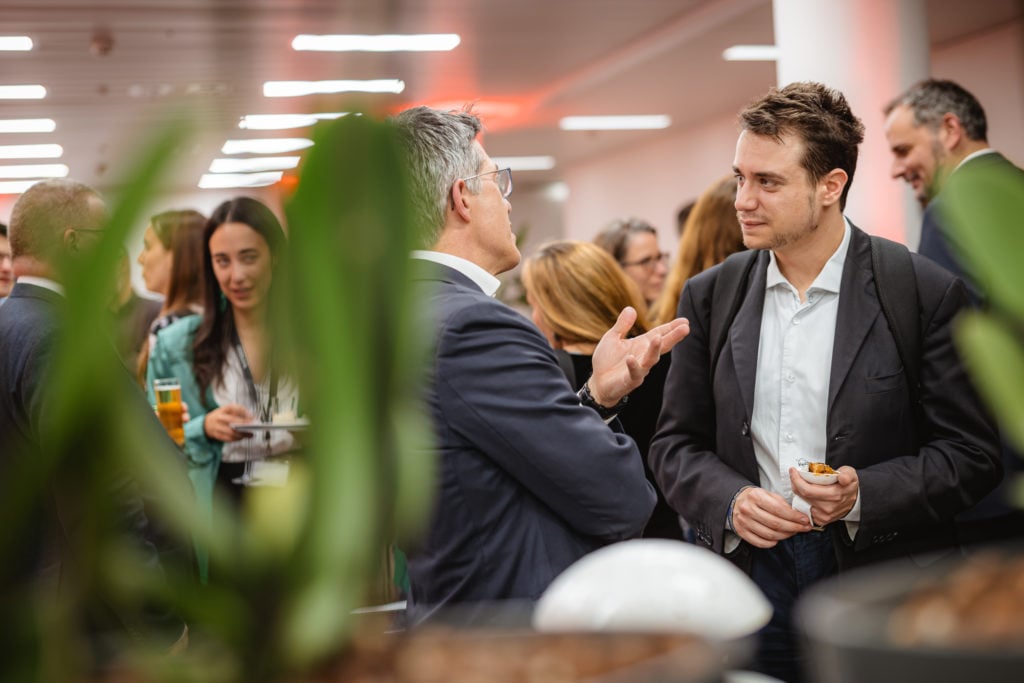 Two men in a business setting engage in conversation at an indoor networking event. One holds a drink, the other a snack. They are surrounded by other attendees. Green plants are in the foreground, and the room is lit with overhead lights and a soft red glow.