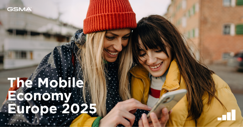 Two women in winter clothing look at a smartphone together, smiling. One wears a red beanie and striped scarf, and the other dons a yellow jacket. They are outside, with blurred buildings in the background. Text reads "GSMA The Mobile Economy Europe 2025" with a signal icon.