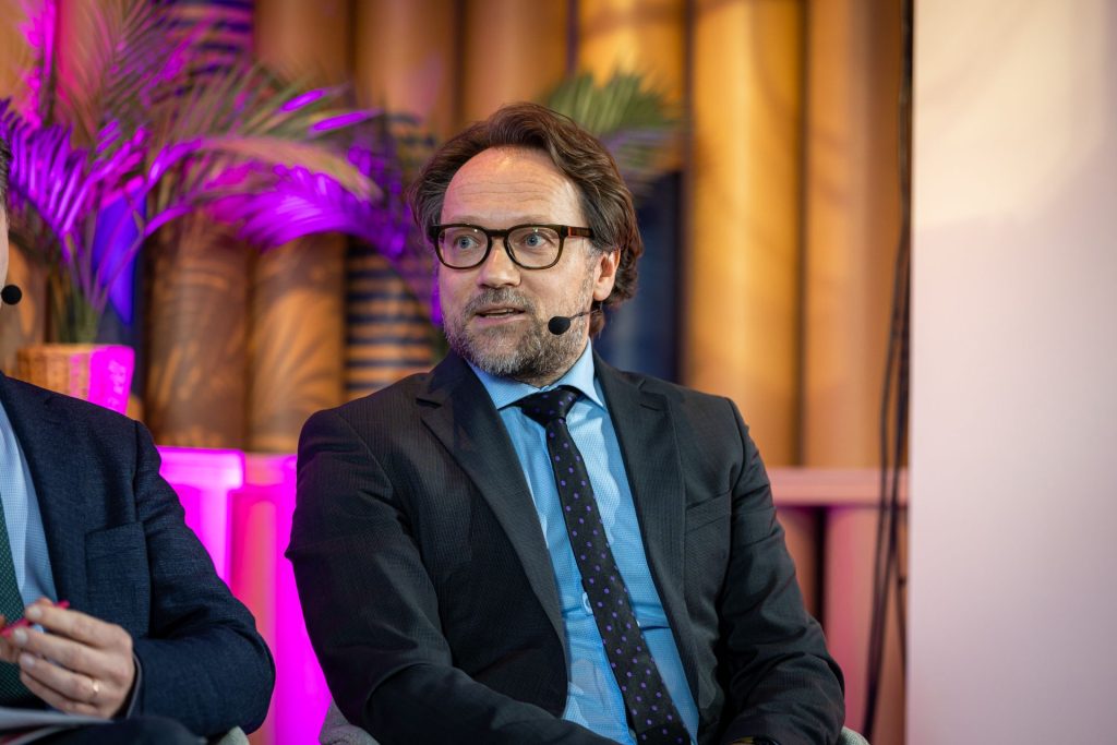 Man with brown hair and glasses, wearing a suit with a polka dot tie, sits on a panel speaking into a headset microphone. Background features tall plants and warm lighting with a gradient of pink to neutral tones.