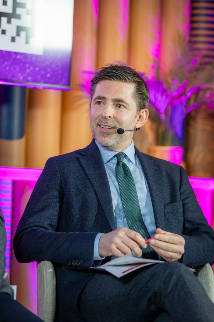 A man in a suit sits on a stage, holding a booklet and pen. He is wearing a dark blue suit, light blue shirt, and green tie. A headset microphone is attached. The background is colorful, with pink lighting and wooden accents. A potted plant and part of a screen are visible.