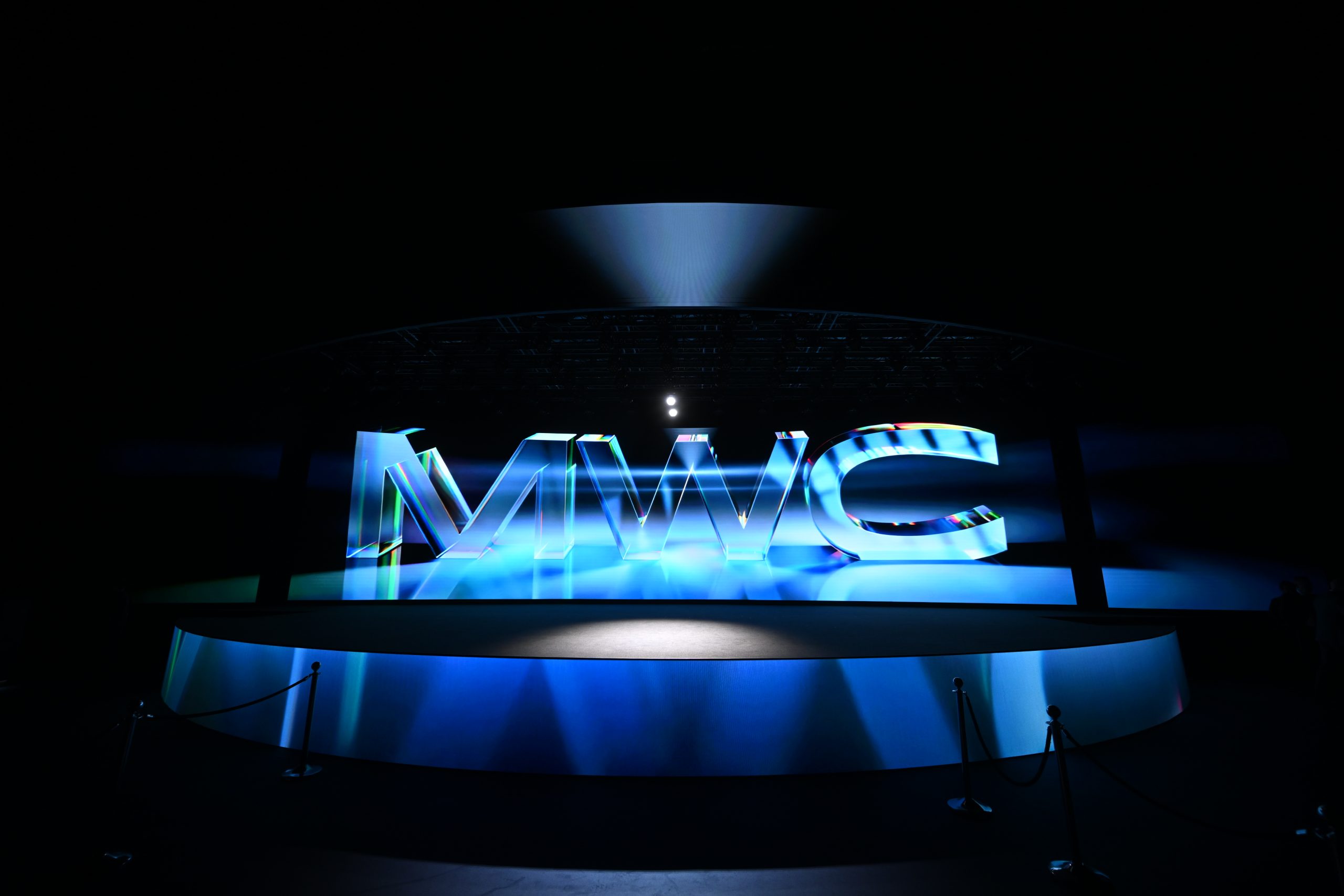 Large, illuminated 3D letters "MWC" stand on a circular stage, surrounded by blue lighting and spotlights, in a dark exhibition space with roped barriers in the foreground.