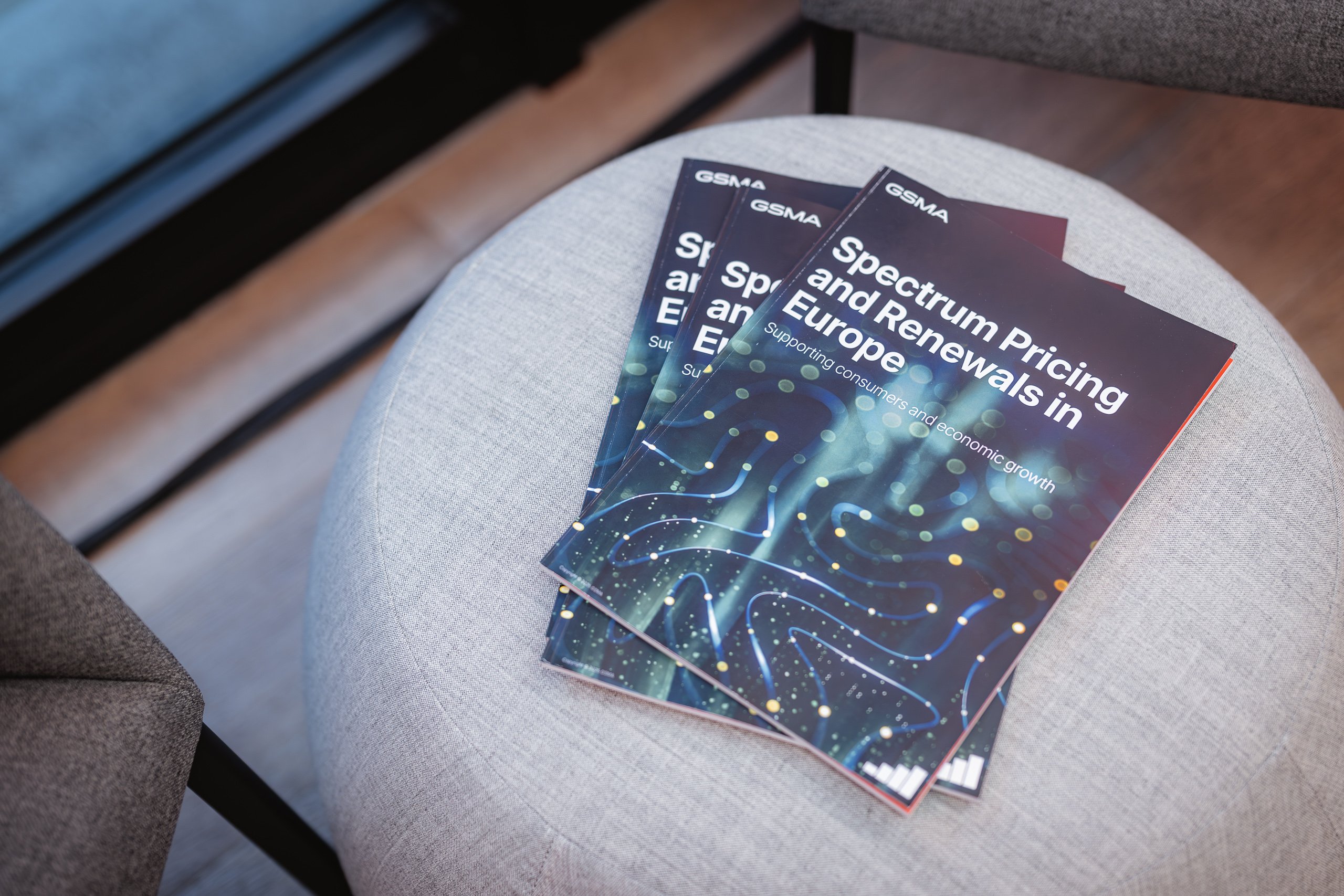 Three copies of a report titled "Spectrum Pricing and Renewals in Europe" rest on a light gray round chair. The cover features abstract blue and green lights. The reports are published by GSMA and subtitled “Supporting Connectivity and Competition.”.