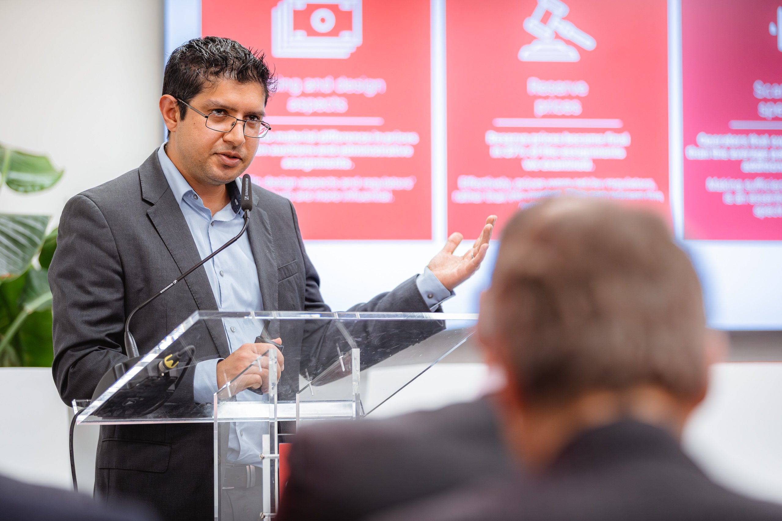 A man in a suit speaks at a clear podium with a microphone, gesturing with one hand. He stands in front of large red and white presentation slides. Audience members are visible in the foreground, seen from behind. A green plant is in the background.