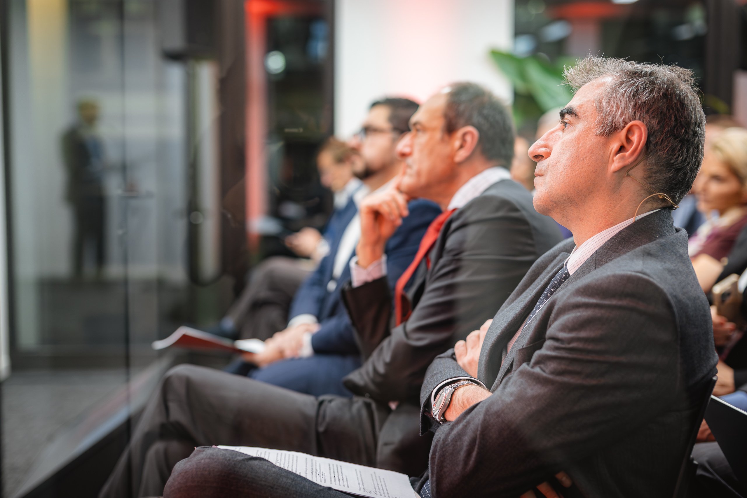 Several people in business attire sit in a row at a conference or seminar, attentively listening. The man in the foreground wears a gray suit and tie, holding papers. The background is softly blurred, with others also appearing focused.