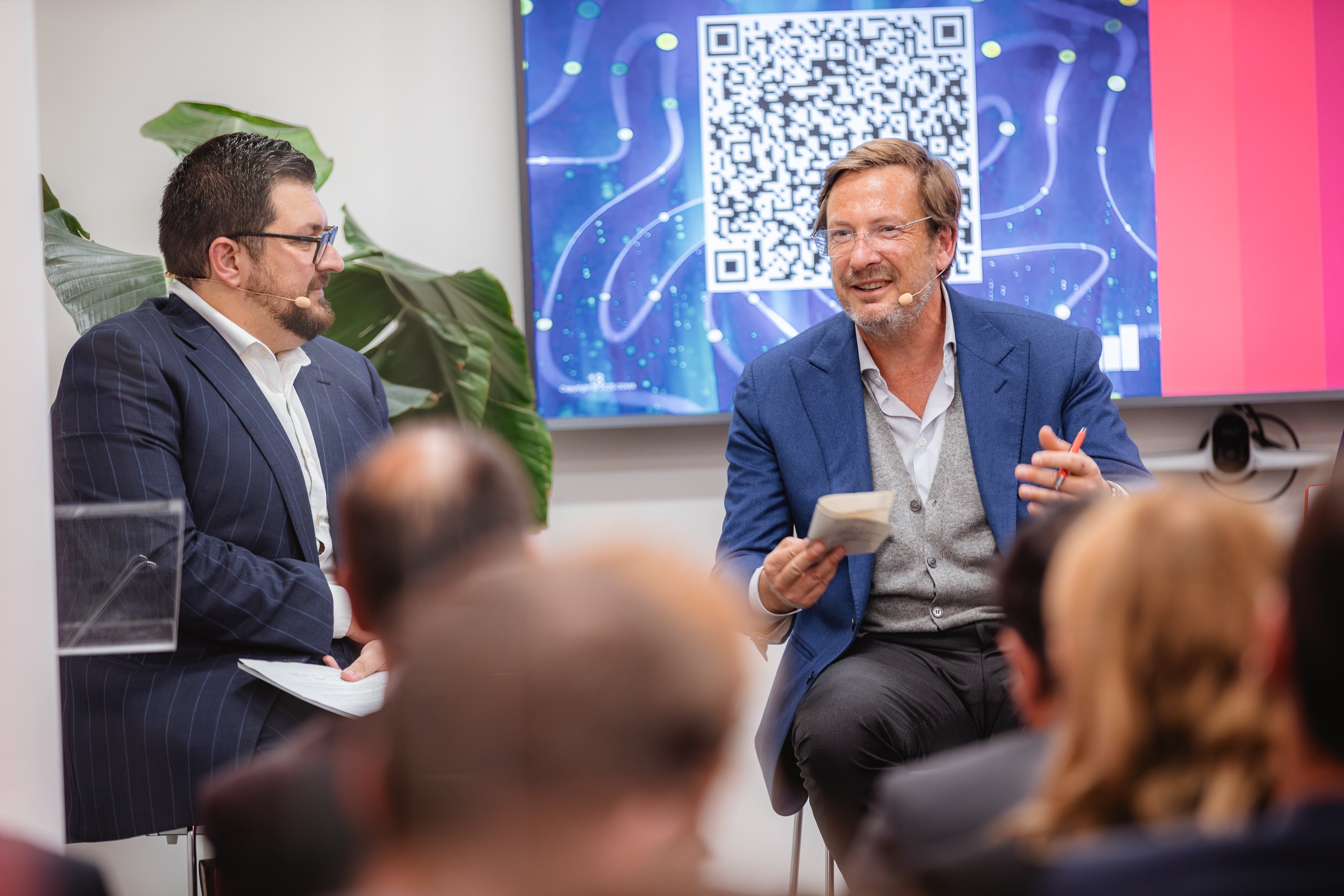 Two men in suits sit and speak on stage at an indoor event, both wearing microphones. One is holding a notepad, gesturing as he talks. A QR code and digital graphics are displayed on a screen behind them. Audience members are visible in the foreground.