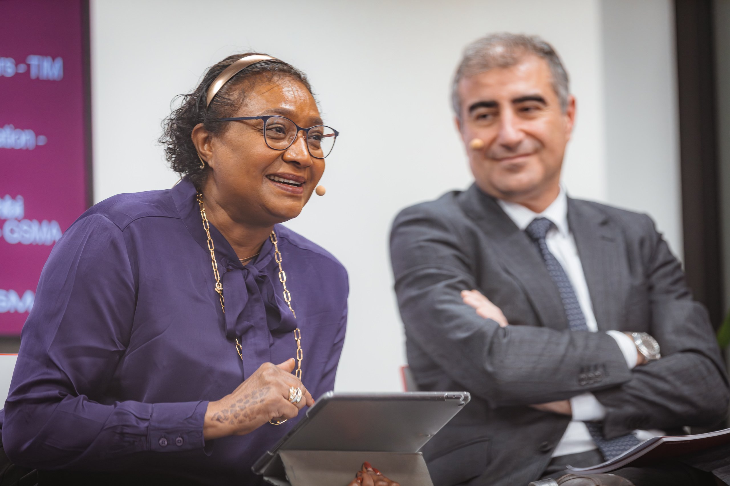 A woman in glasses and a purple blouse speaks while holding a tablet, smiling, with a headset microphone. A man in a suit with crossed arms and a headset microphone sits beside her, looking on. Both appear engaged in a panel discussion.