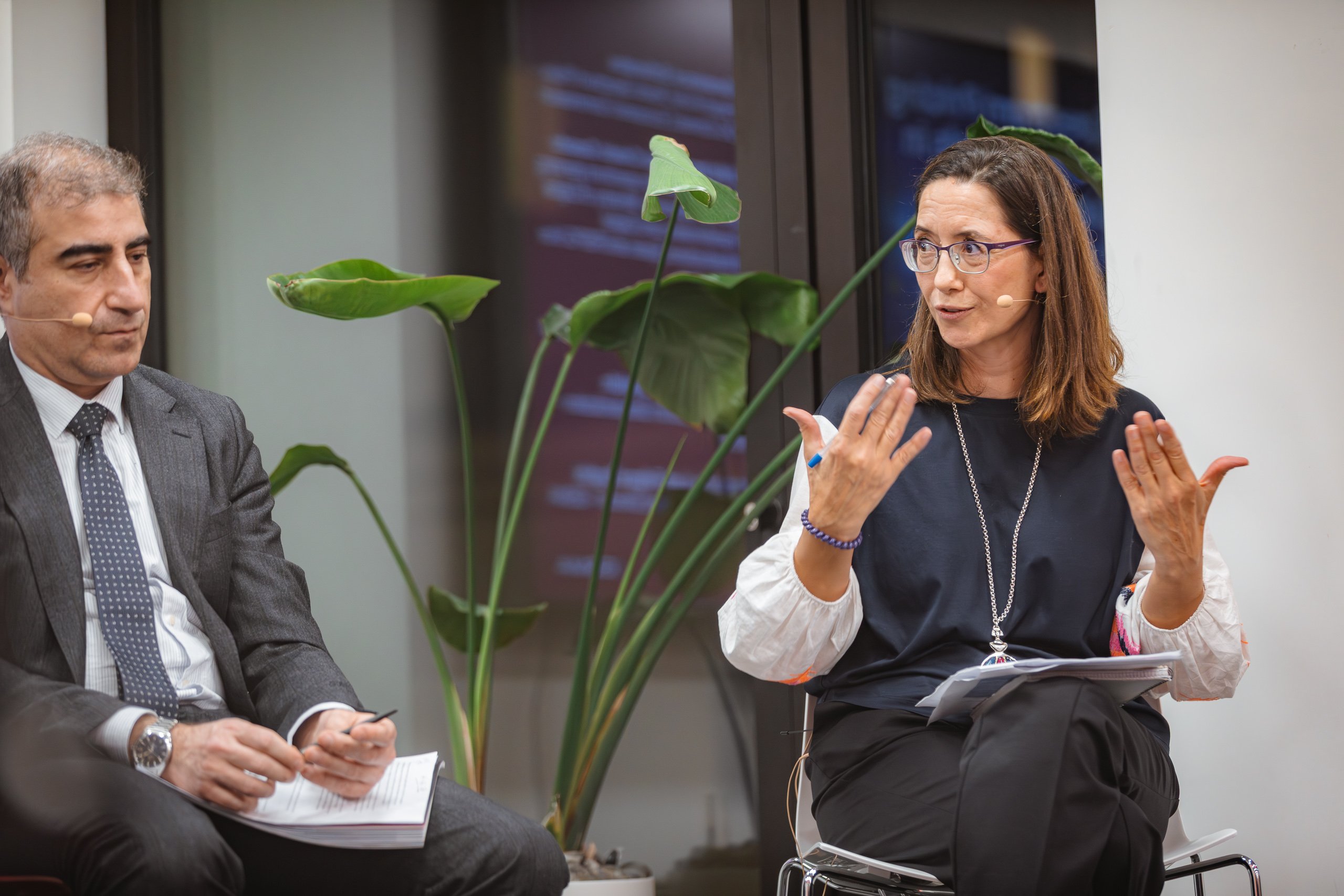 A man in a suit and a woman in business casual clothes sit and talk indoors. Both wear headset microphones. The woman gestures while speaking, and the man listens, holding a pen and notebook. A large green plant and glass window are visible behind them.