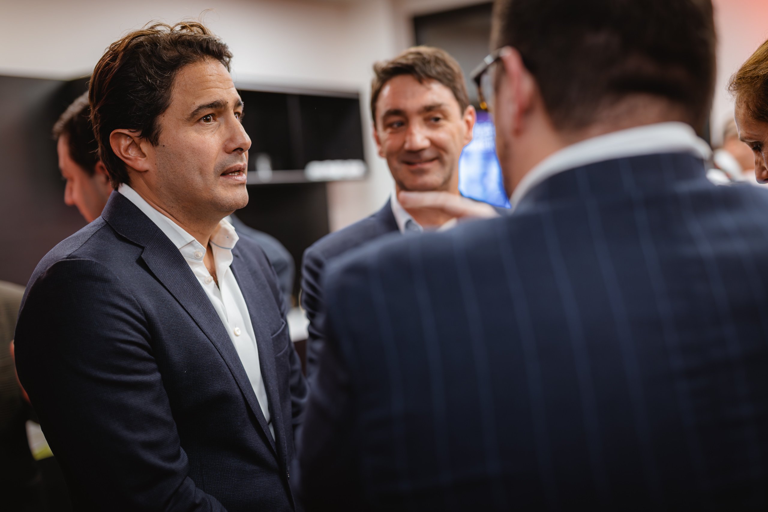 A man in a navy suit stands indoors, talking and listening to others in business attire. Another man in a suit stands nearby, smiling. The setting appears professional, with blurred figures and office-like surroundings in the background.