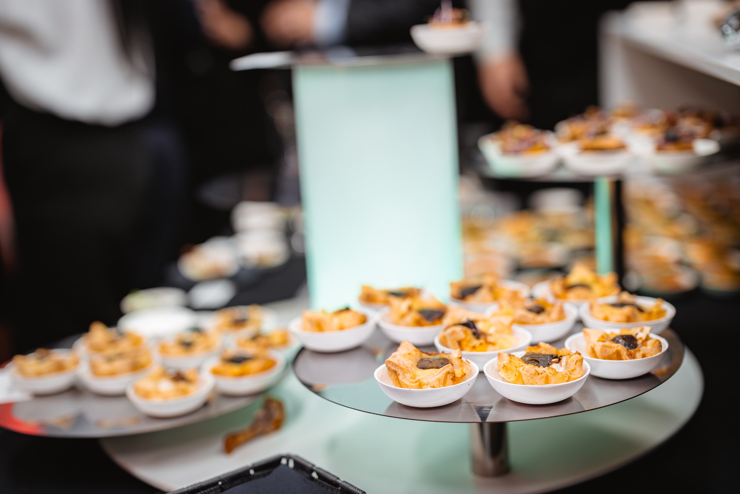 A display of small white bowls filled with bite-sized appetizers, possibly pasta or pastry with garnish, arranged on a circular tiered stand. The background is blurred with people standing, suggesting a catered event or buffet.