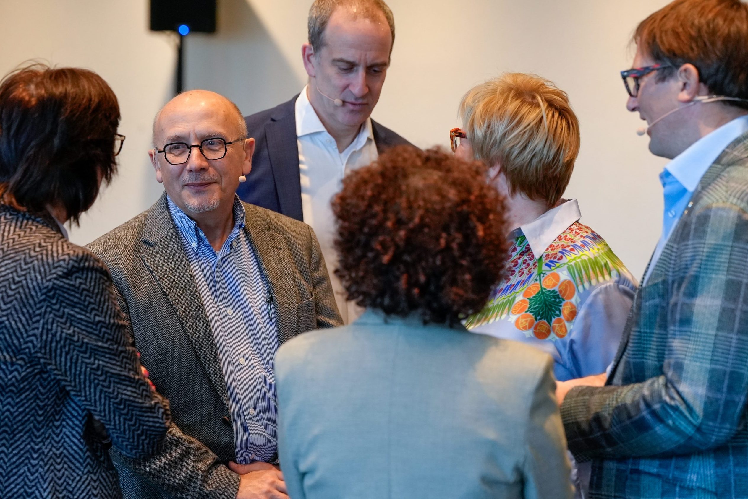Six adults, four facing away from the camera, stand in a close group, engaged in conversation. One man in glasses and a grey blazer smiles. The group is dressed in business or smart casual attire, standing indoors against a plain background.