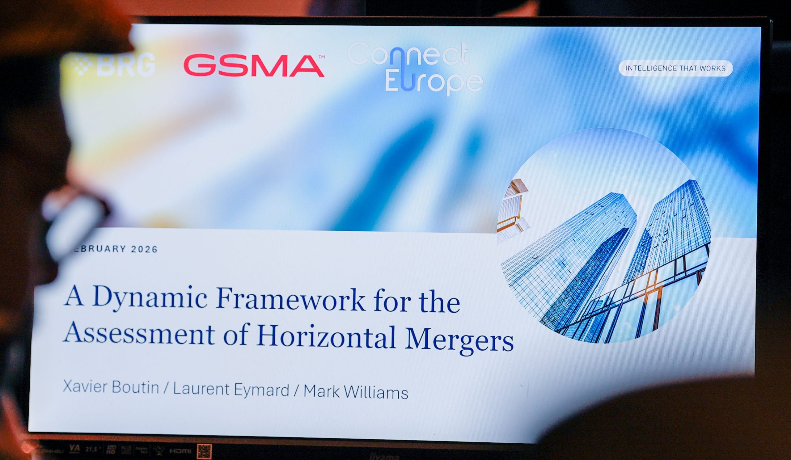 A computer screen displays a conference slide titled “A Dynamic Framework for the Assessment of Horizontal Mergers” with authors Xavier Boutin, Laurent Eymard, and Mark Williams. The slide has logos for BRG and GSMA, and shows modern glass buildings inside a circular graphic.