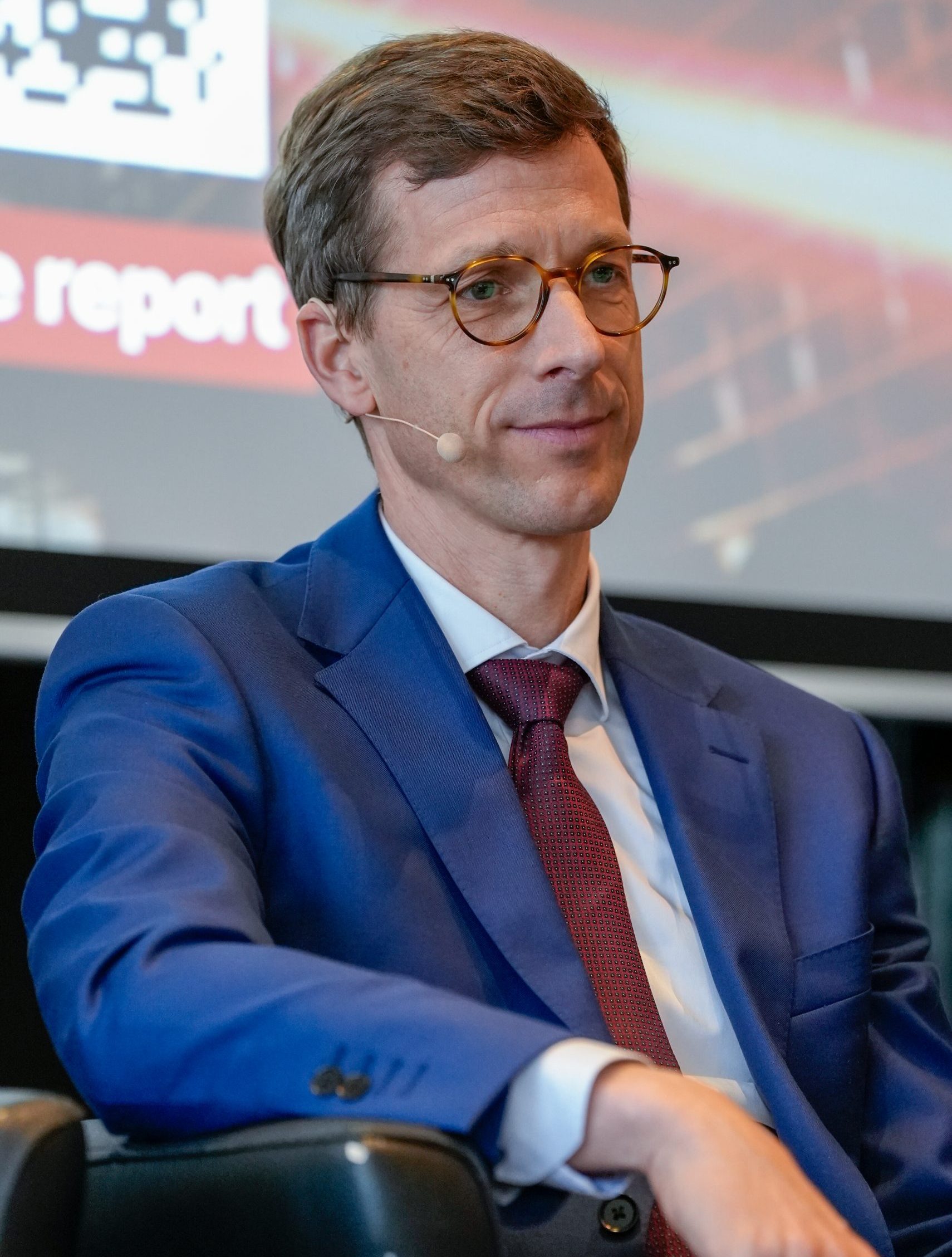 A man in a blue suit, red tie, and glasses sits on a chair, wearing a headset microphone. He has short brown hair and a neutral expression. In the background, there is a blurred screen with red, white, and black graphics and partial text.