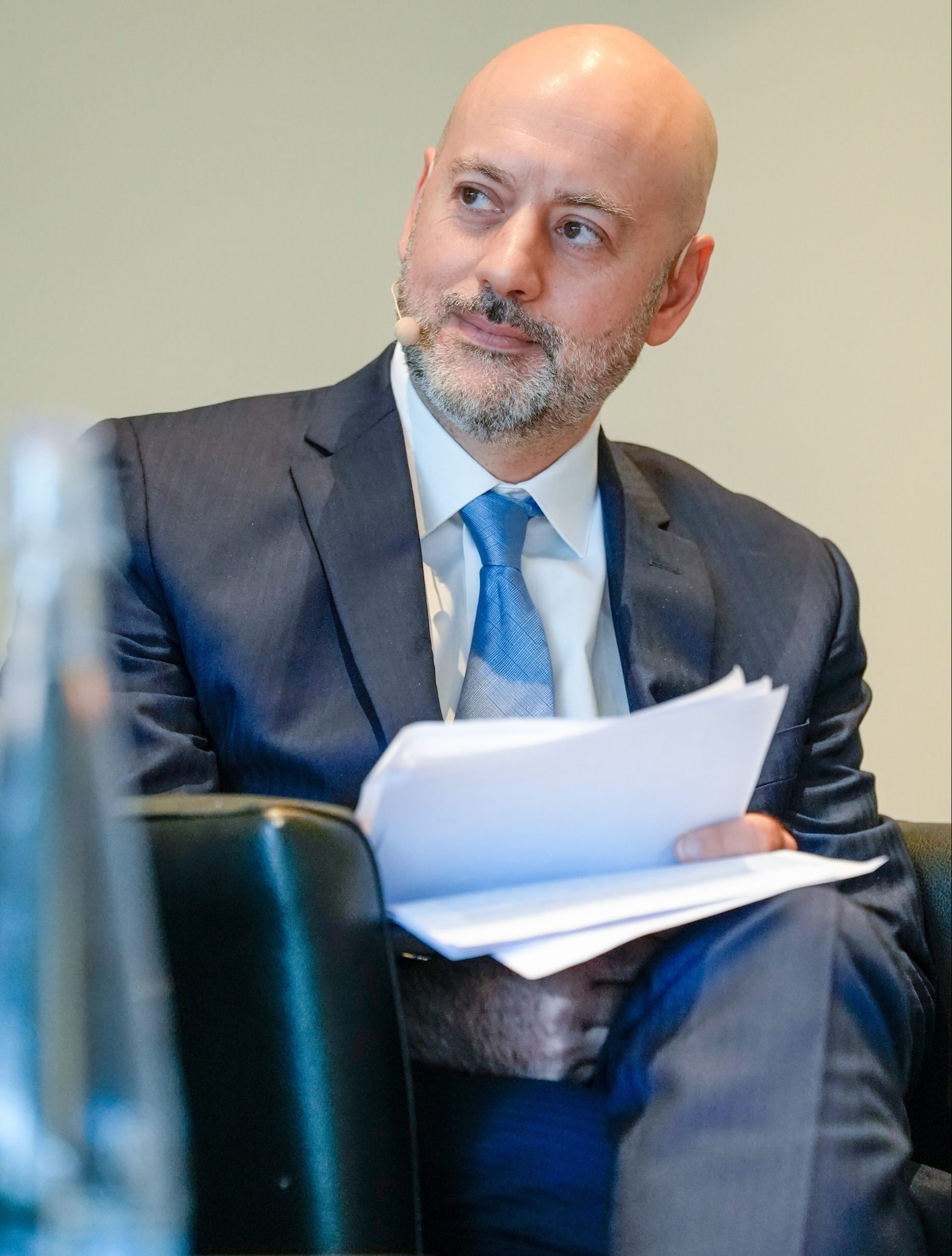 A bald man with a short beard, wearing a navy blue suit and a blue tie, sits holding papers on his lap. He appears to be at a formal event, looking to the side while listening attentively. A blurred glass bottle is visible in the foreground.