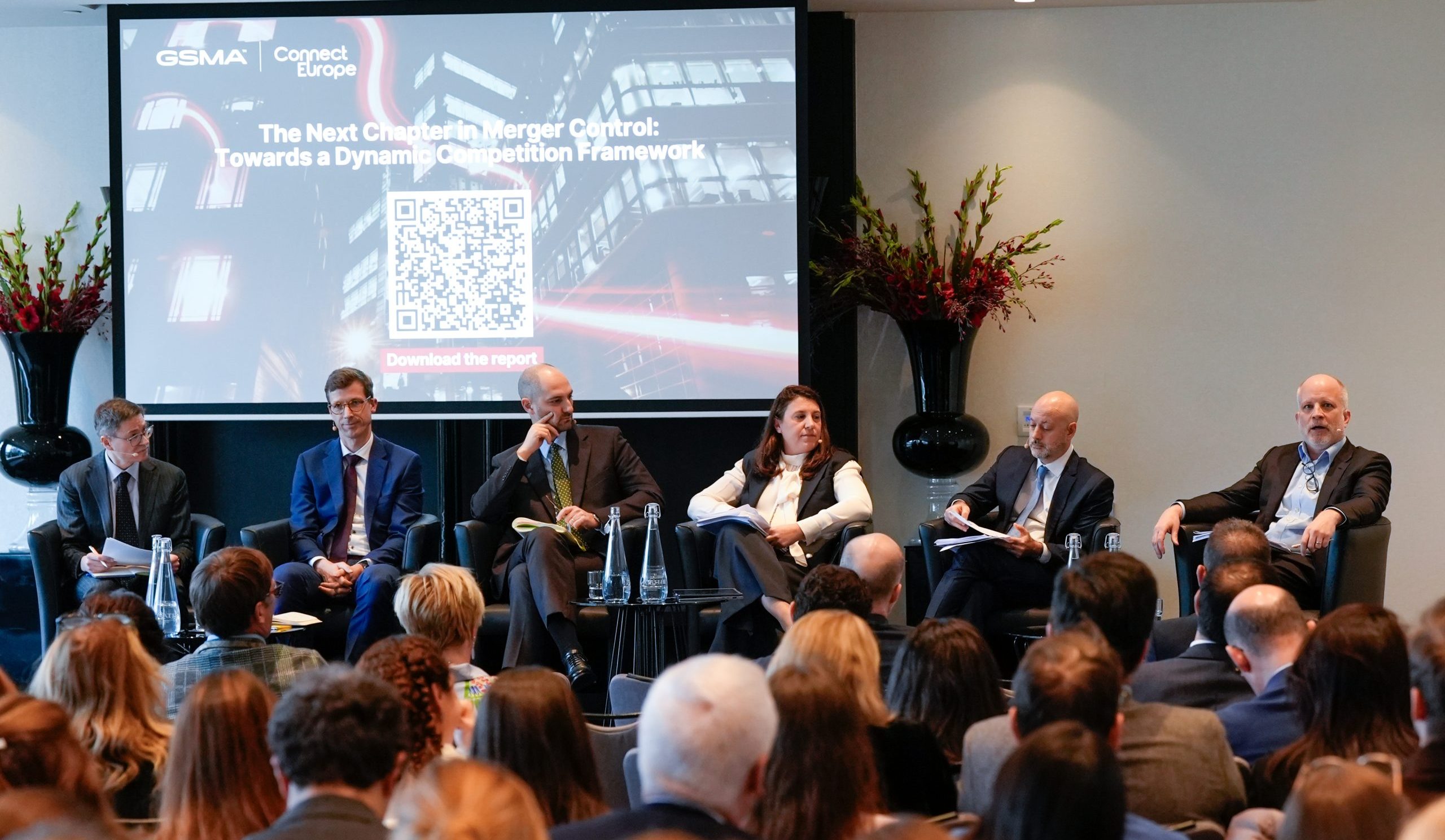 Six panelists in business attire sit on stage in front of an audience. Behind them is a large screen with the text: “The Next Chapter in Merger Control: Towards a Dynamic Competition Framework” and a prominent QR code, flanked by tall floral arrangements.