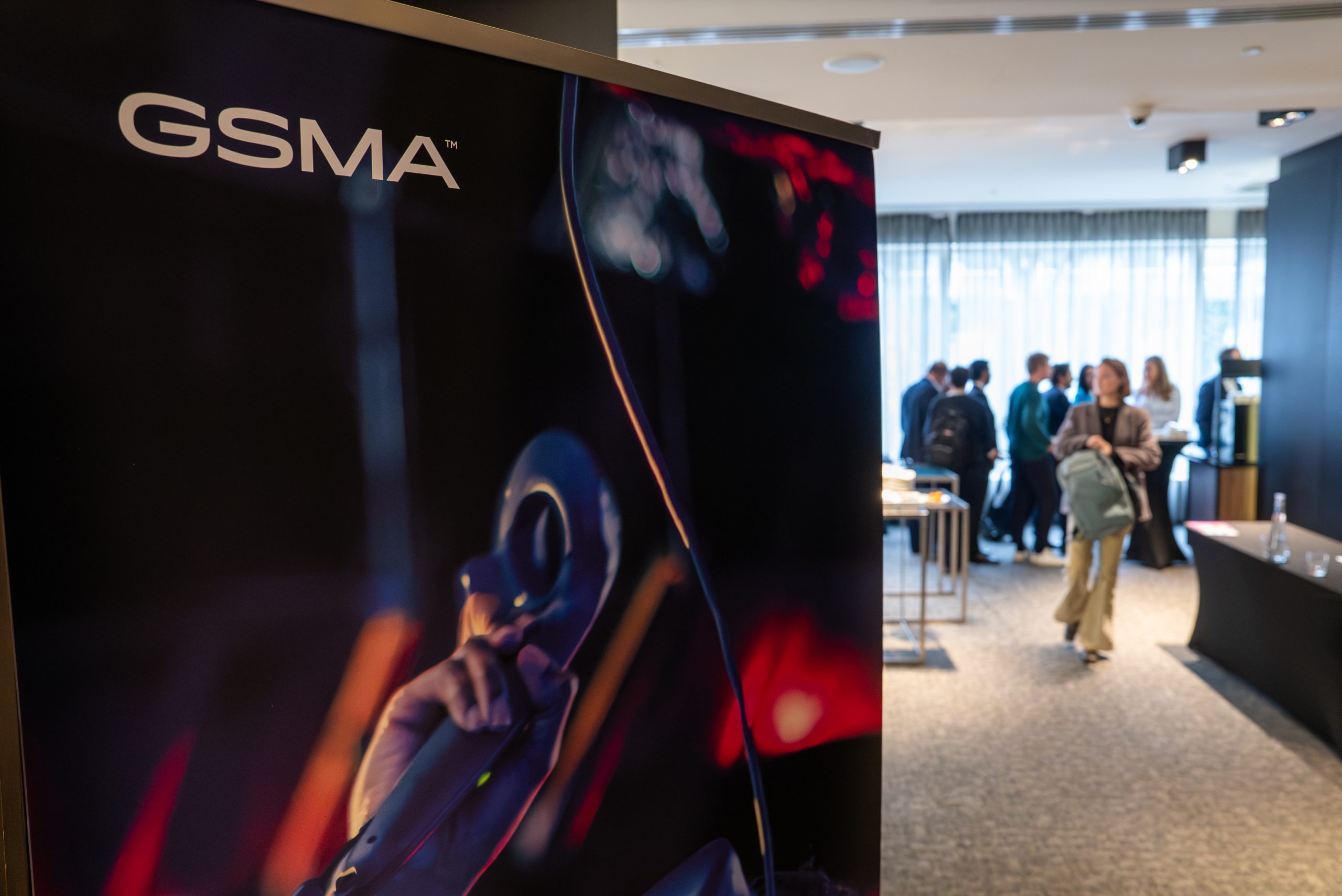 A GSMA banner featuring a close-up of a hand holding a microphone stands in the foreground; in the background, people are gathered and conversing in a brightly lit, modern event space with large windows.