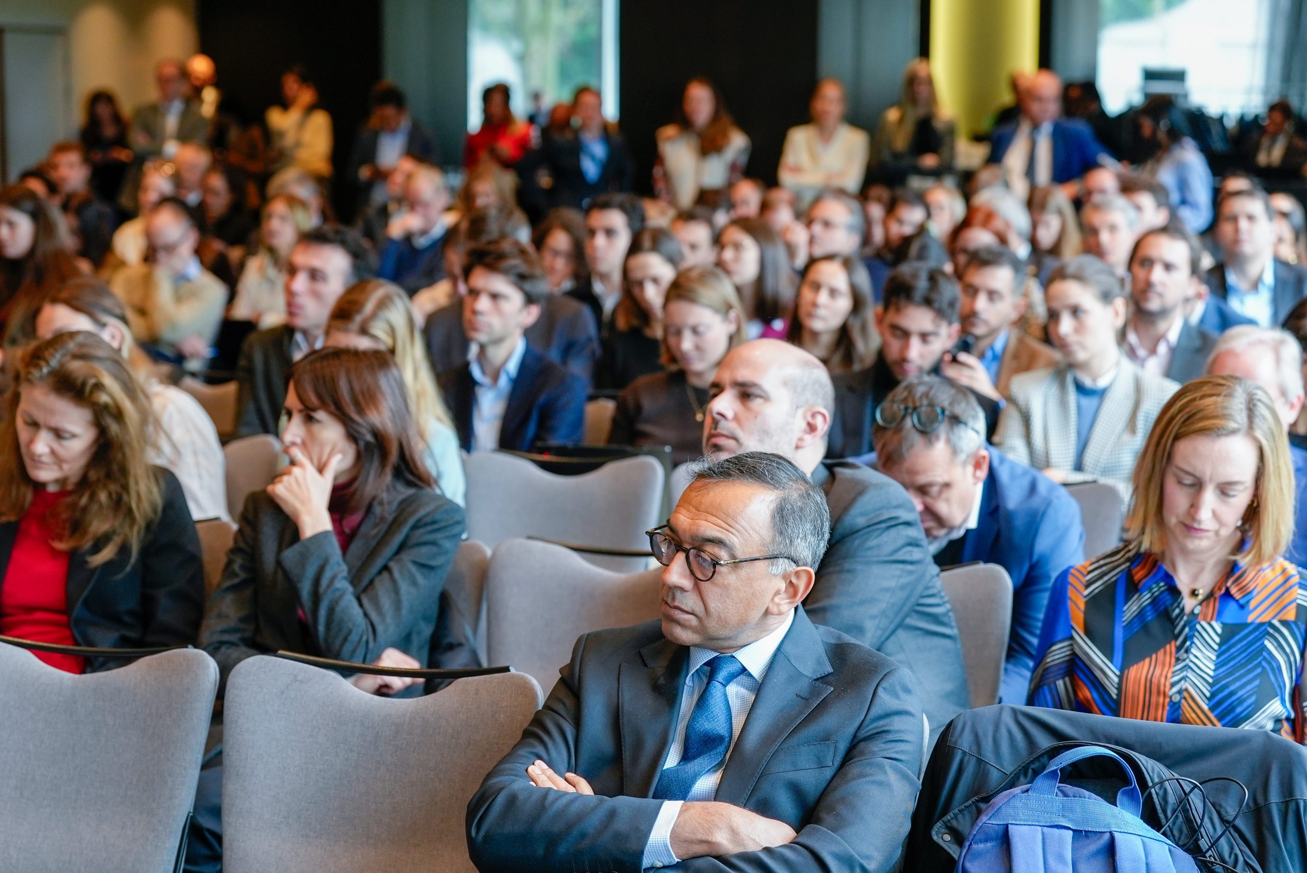 A large group of people sit closely together in rows at a conference or seminar, listening attentively. Most people wear business or smart casual attire. Expressions range from focused to neutral. The background is slightly blurred, indicating a packed room.