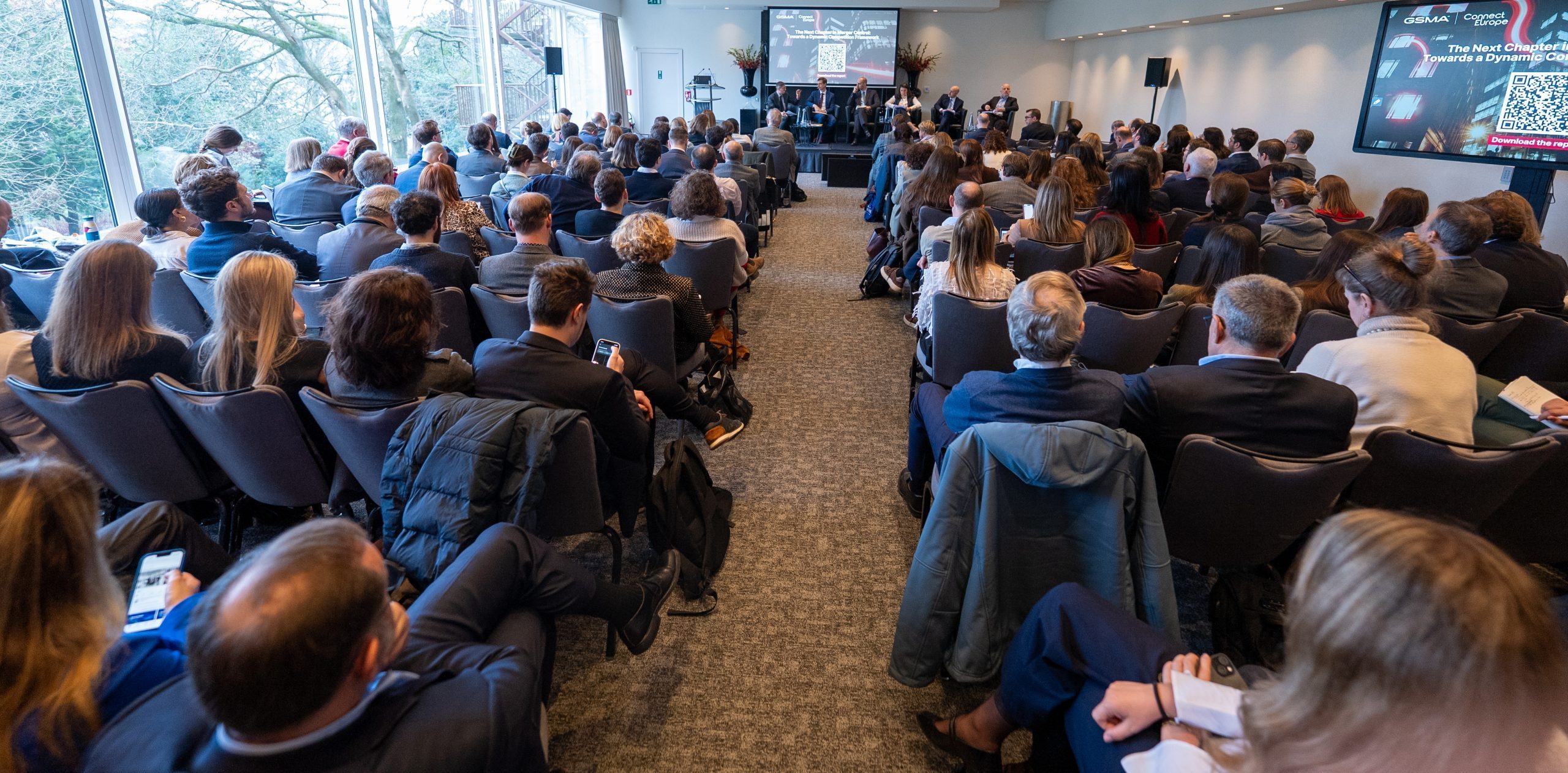 A large group of people in business attire are seated in rows facing a stage, where speakers present from a podium. The room is modern with floor-to-ceiling windows on one side, showing trees outside. A screen displays presentation slides above the speakers.
