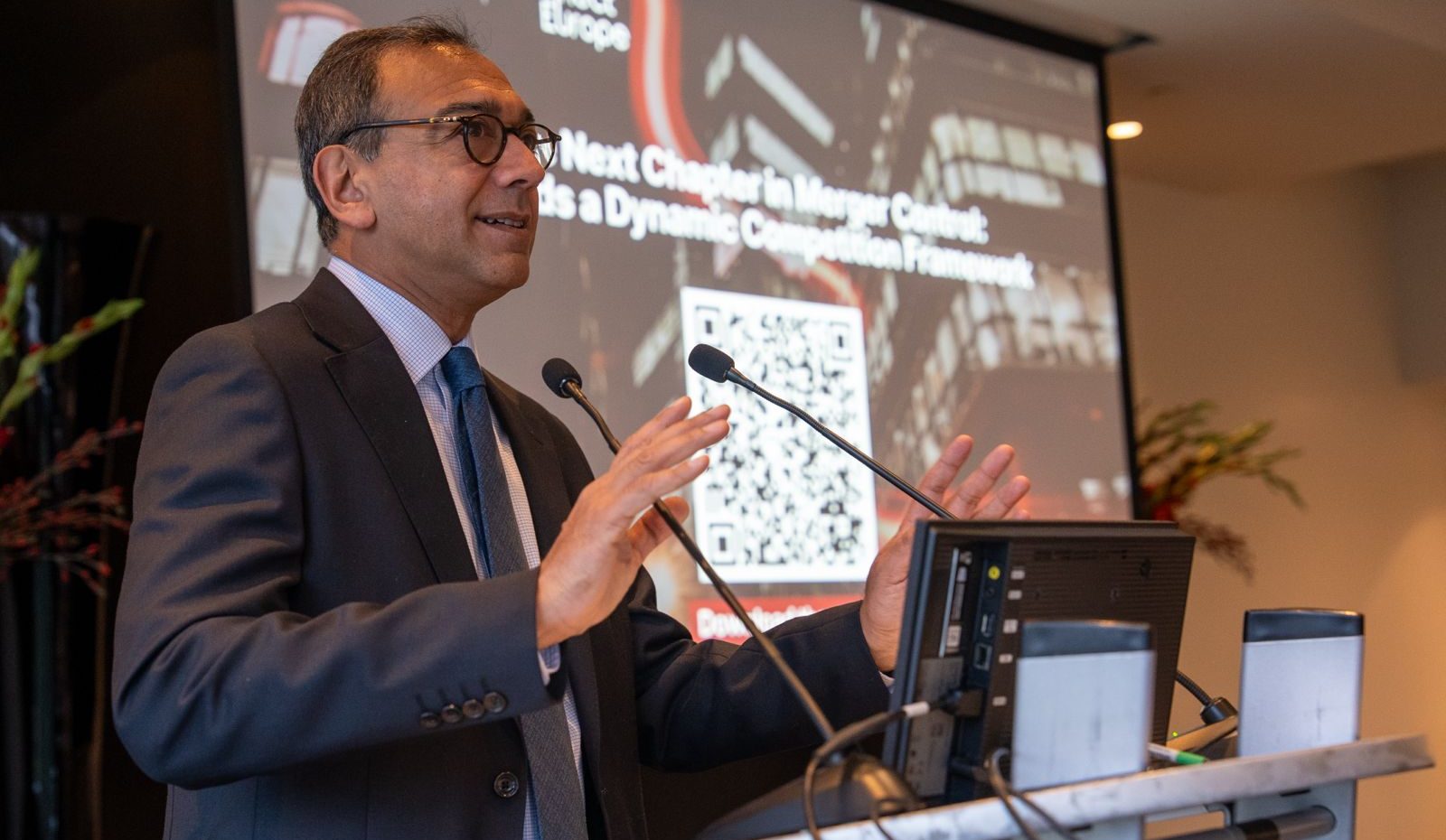 A man in a suit and glasses speaks at a podium with microphones. Behind him is a screen displaying a QR code, text, and cityscape images. The text reads, “Next Chapter in Merger Control: Towards a Dynamic Competition Framework.” Plants are visible in the background.
