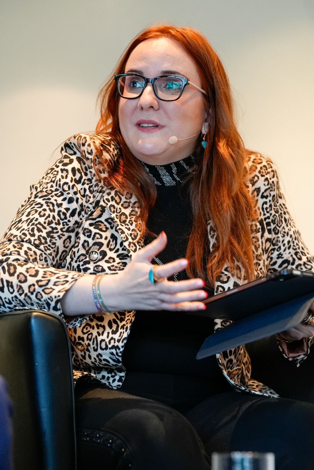 A woman with long red hair, glasses, and a headset mic sits in a black chair. She wears a leopard print jacket over a black outfit, gestures with one hand, and holds a tablet in the other, appearing engaged in conversation.