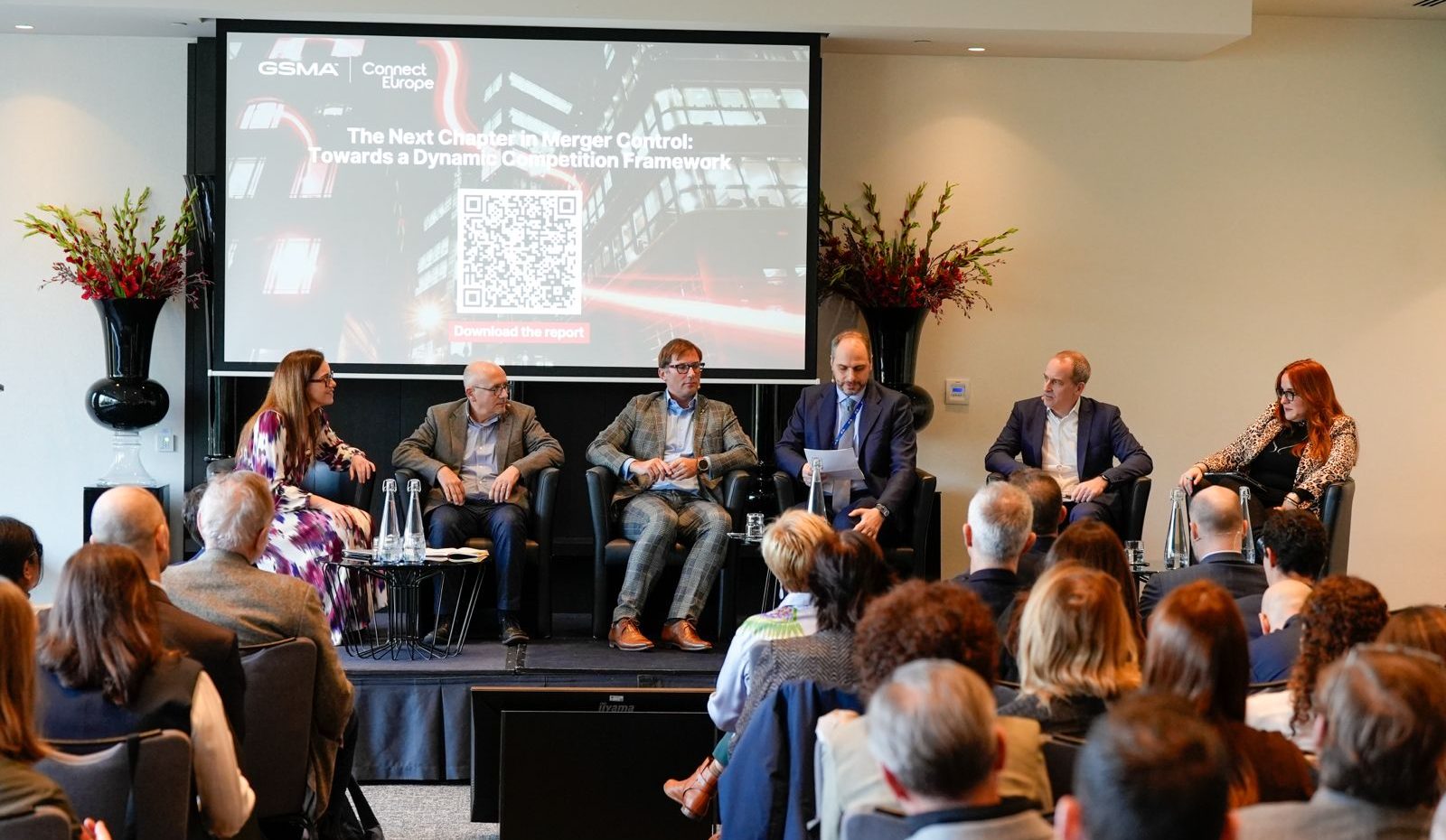 A panel of six people, four men and two women, sit on a stage facing an audience, discussing in front of a screen displaying the GSMA logo and event title. The room is modern, with flower arrangements and a QR code on the presentation slide.