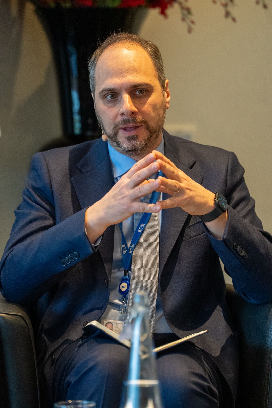 A man with short dark hair and a trimmed beard sits in a chair, wearing a navy suit, light shirt, and tie. He clasps his hands in front of him, appearing attentive and engaged. He wears a headset and conference lanyard, with a blurred background.