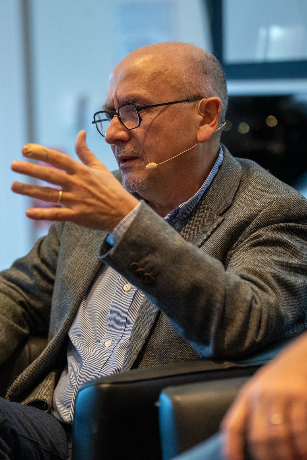 A bald man wearing glasses, a gray blazer, and a striped shirt sits in a black chair, speaking with his left hand raised. He has a thin microphone headset on his cheek and appears engaged in conversation during an indoor event or discussion.