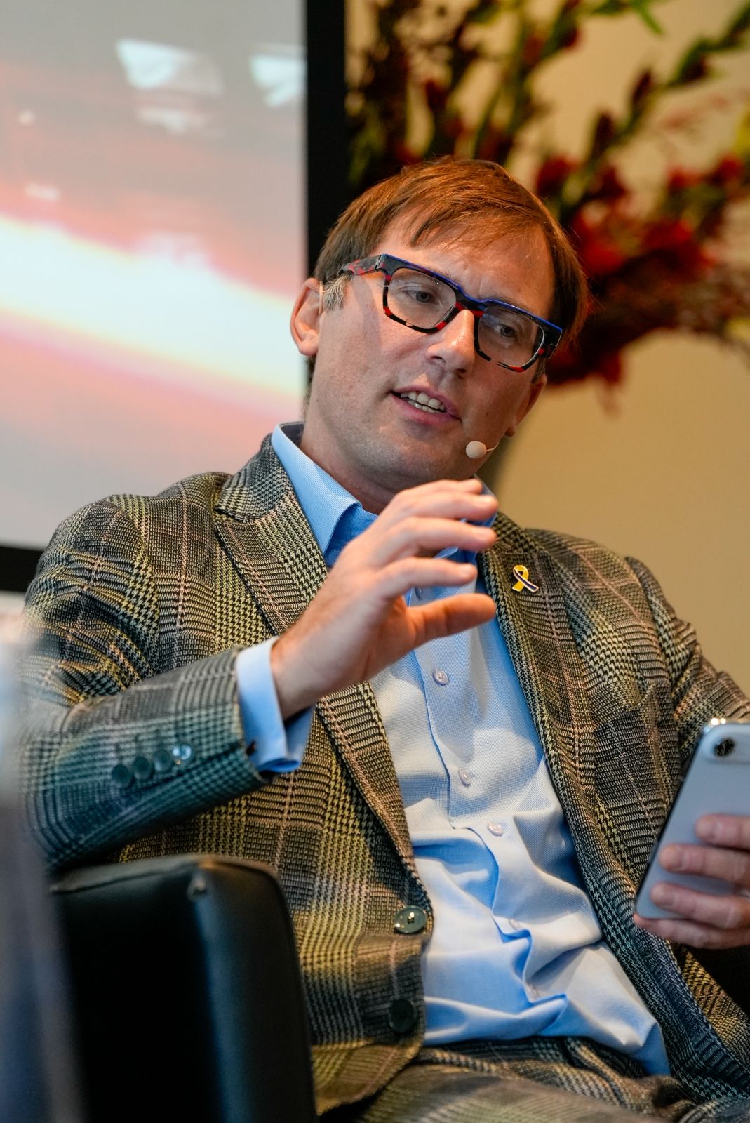 A man with light brown hair, glasses, and a plaid suit jacket is seated, holding a smartphone and gesturing with his other hand. He wears a blue shirt and a pin, and has a headset mic. A blurred screen and red flowers are visible in the background.