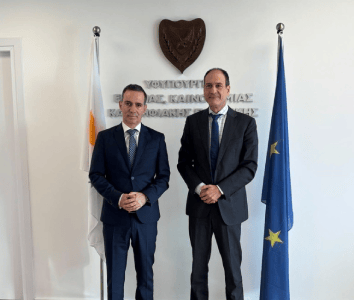 Two men in suits stand between Cyprus and EU flags, with a wall plaque and text in the background.