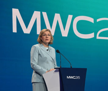 A woman in a light blue suit speaks at a lectern with "MWC26" displayed on the screen behind her.