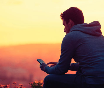 Man in a hoodie sitting outdoors at sunset, looking at and using his mobile phone.