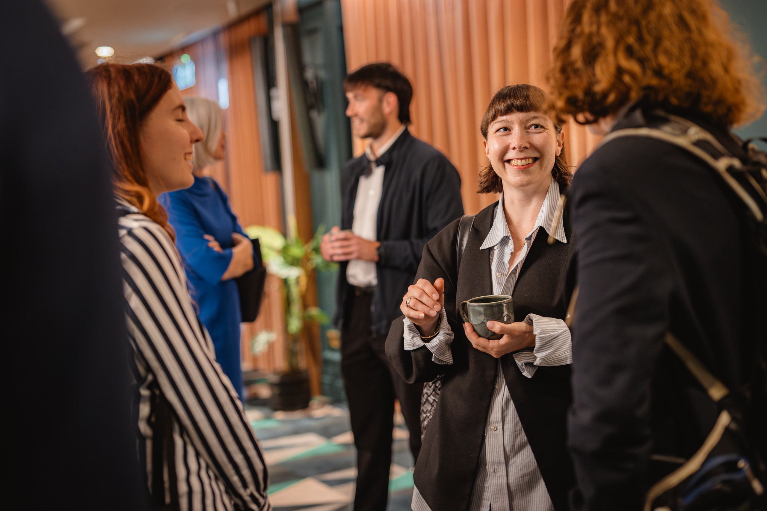 A group of people, both men and women, are standing indoors, chatting and smiling. One woman in focus holds a coffee cup and laughs whilst talking to another person. The setting appears to be informal, possibly a networking event or social gathering.