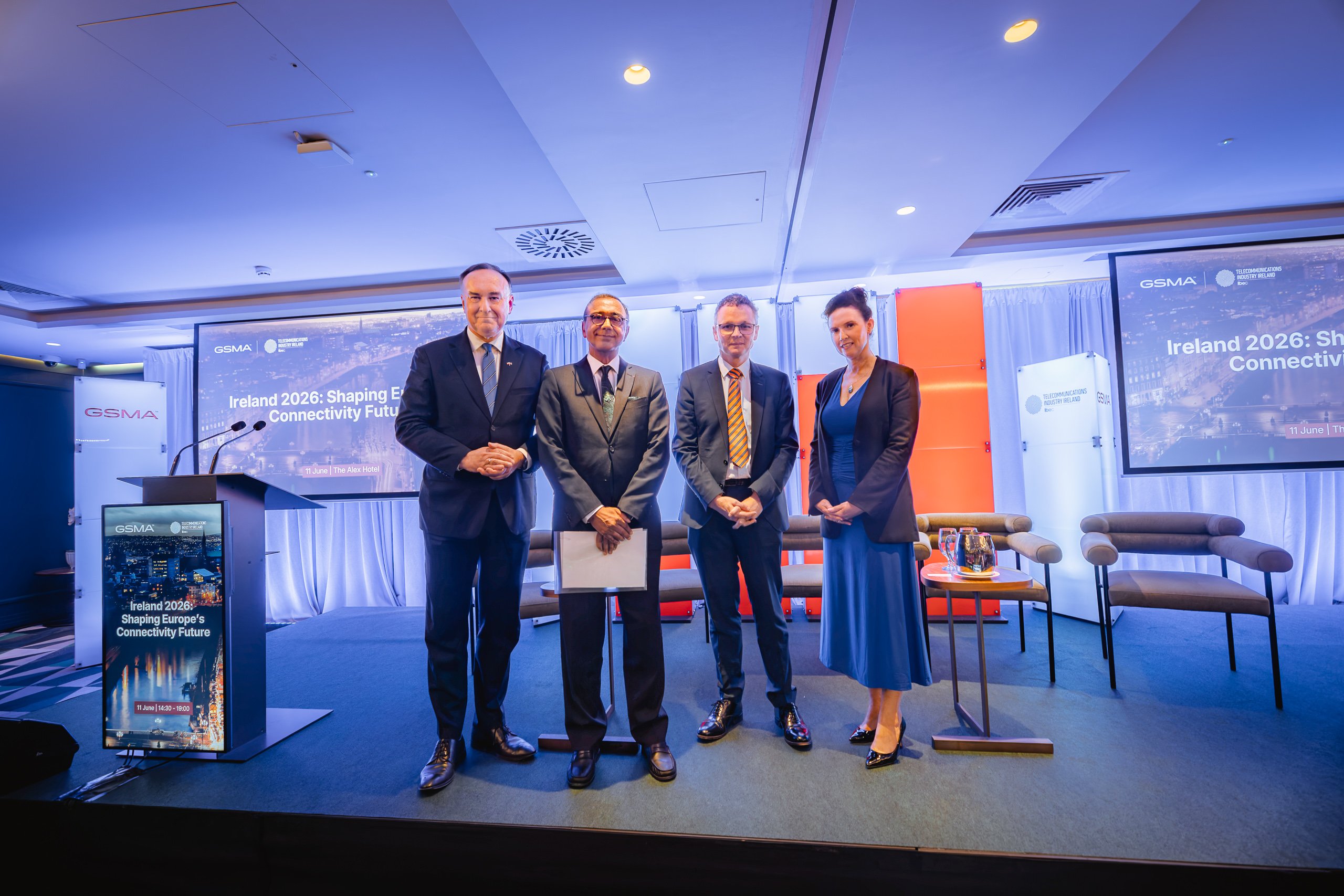 Four professionally dressed individuals (three men, one woman) stand on a stage with an "Ireland 2026: Shaping Europe's Connectivity Future" backdrop. The stage has chairs, a table with water, and blue and orange lighting, creating a formal conference setting.