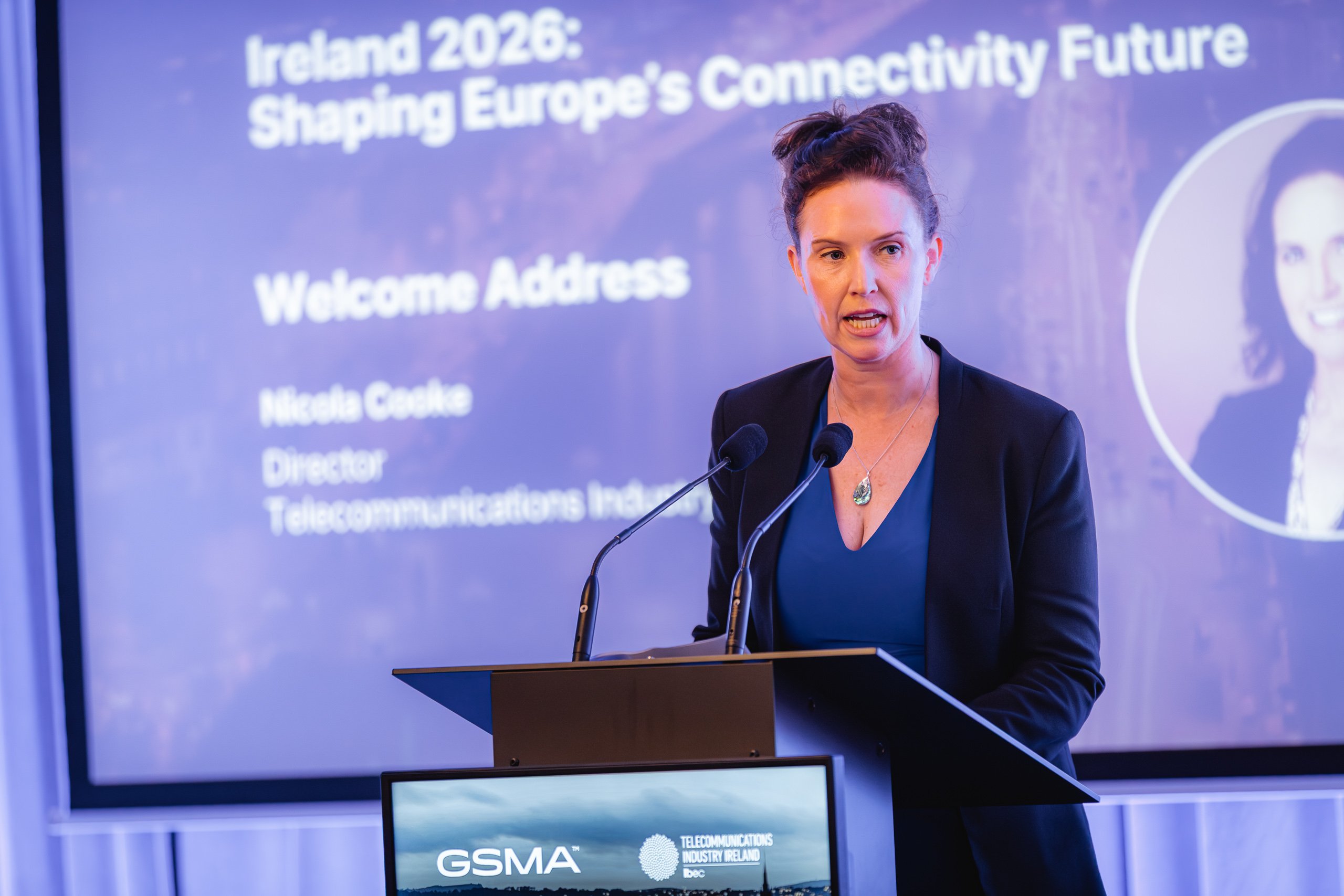 A woman speaks at a lectern with two microphones. Behind her, a large screen displays the text: “Ireland 2026: Shaping Europe’s Connectivity Future” and “Welcome Address – Nicola Cooke, Director, Telecommunications Industry Ireland”.